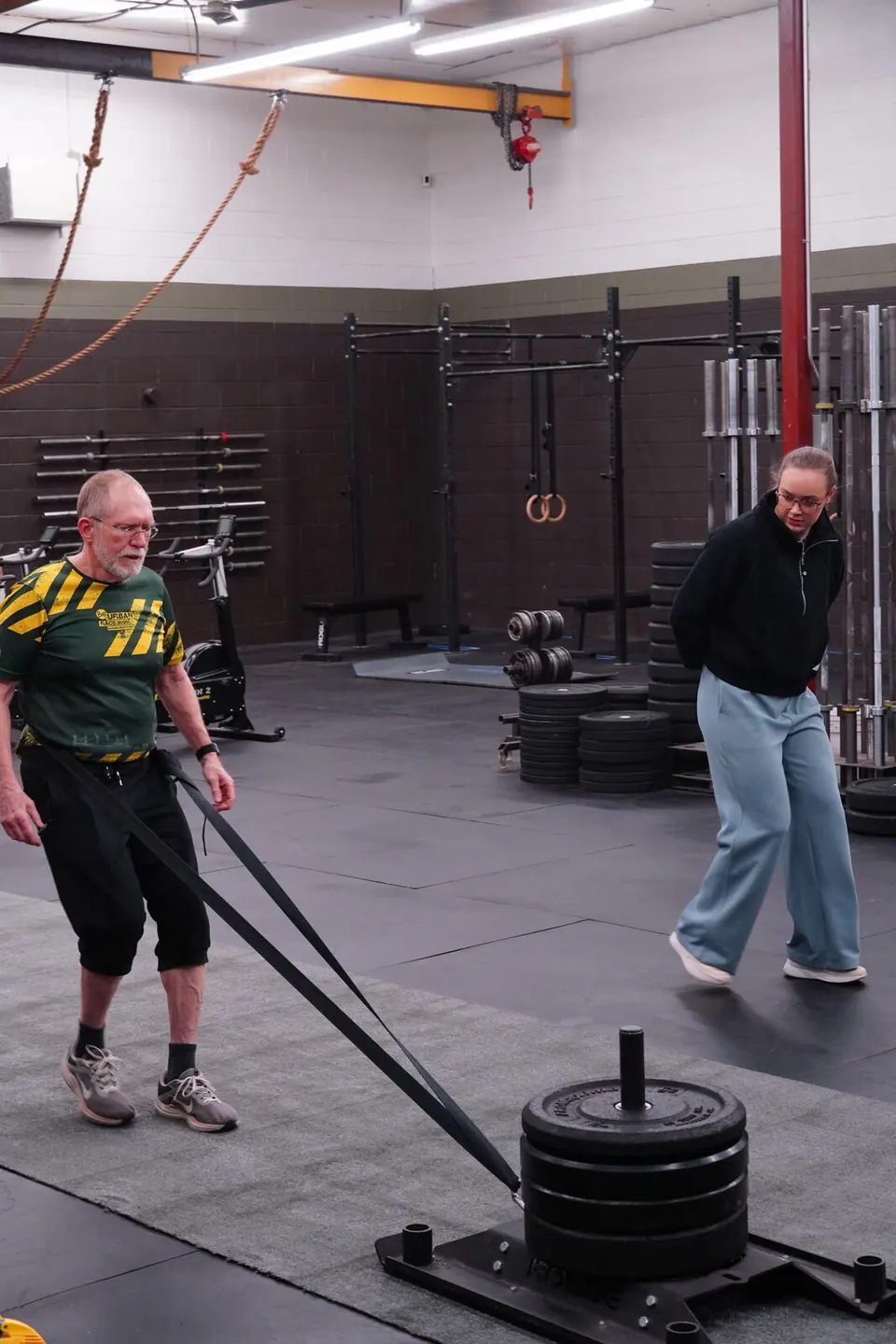 Vitality 55+ member pulling a weighted sled with a strap while a classmate looks on